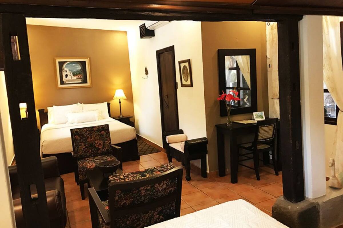 A hotel room with a bed, two chairs, a desk, a mirror, and a painting on the wall.