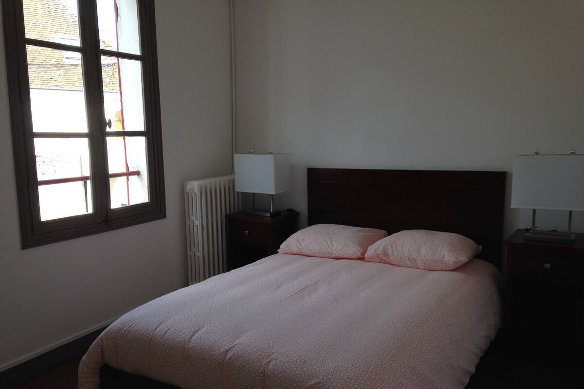 A bedroom with a bed, two bedside tables with lamps, a radiator, and a window with blinds.