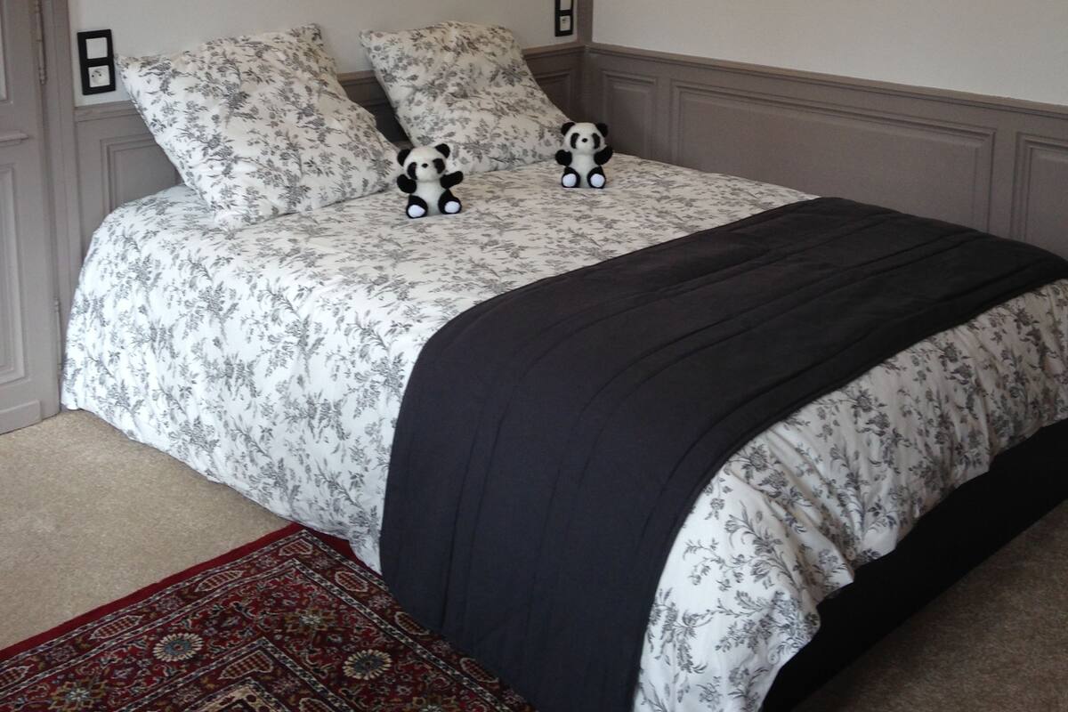 A bedroom with a floral bedspread, two panda plush toys, a dark throw blanket, and two wall-mounted lamps.