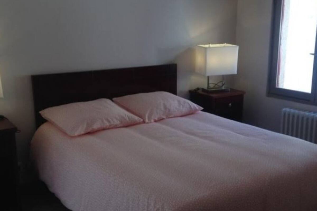 A neatly made bed with a dark headboard and two pillows, a bedside table with a lamp, and a window with daylight coming through.