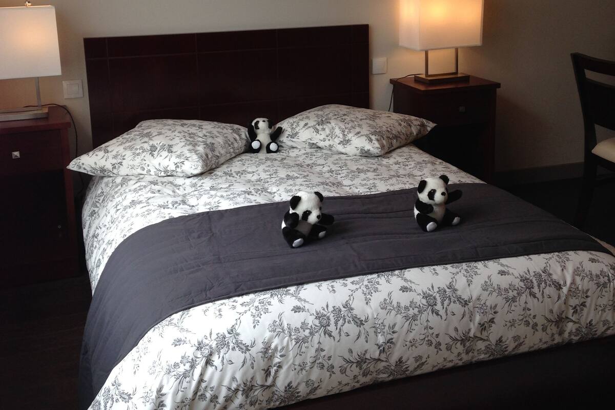 A neatly made bed with floral bedding and two panda plush toys. There are two bedside lamps and a chair visible.