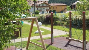 Un parque infantil con columpios y un puente colgante en una zona residencial con casas al fondo.