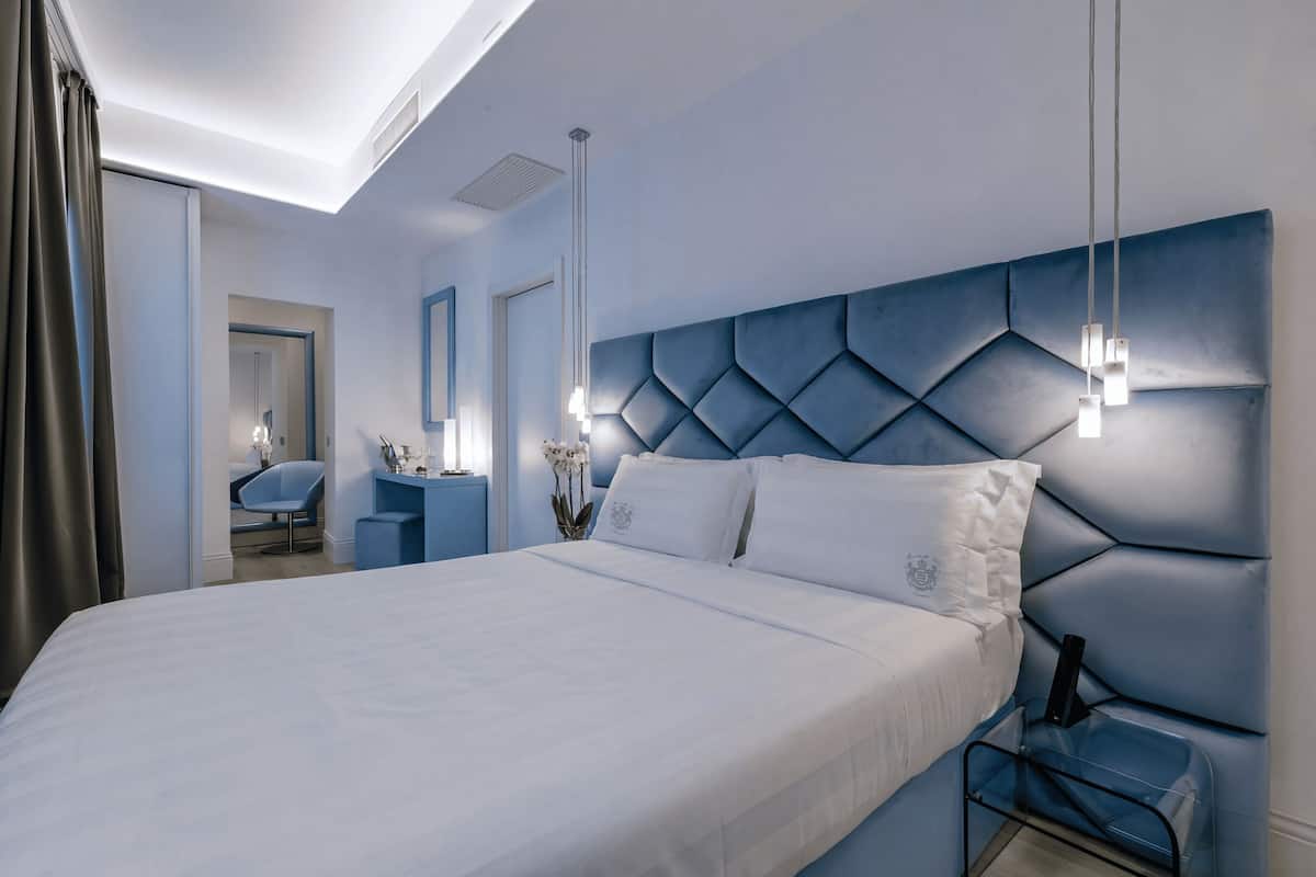 A modern hotel room with a large bed, a blue headboard, a desk with a chair, and a mirror.