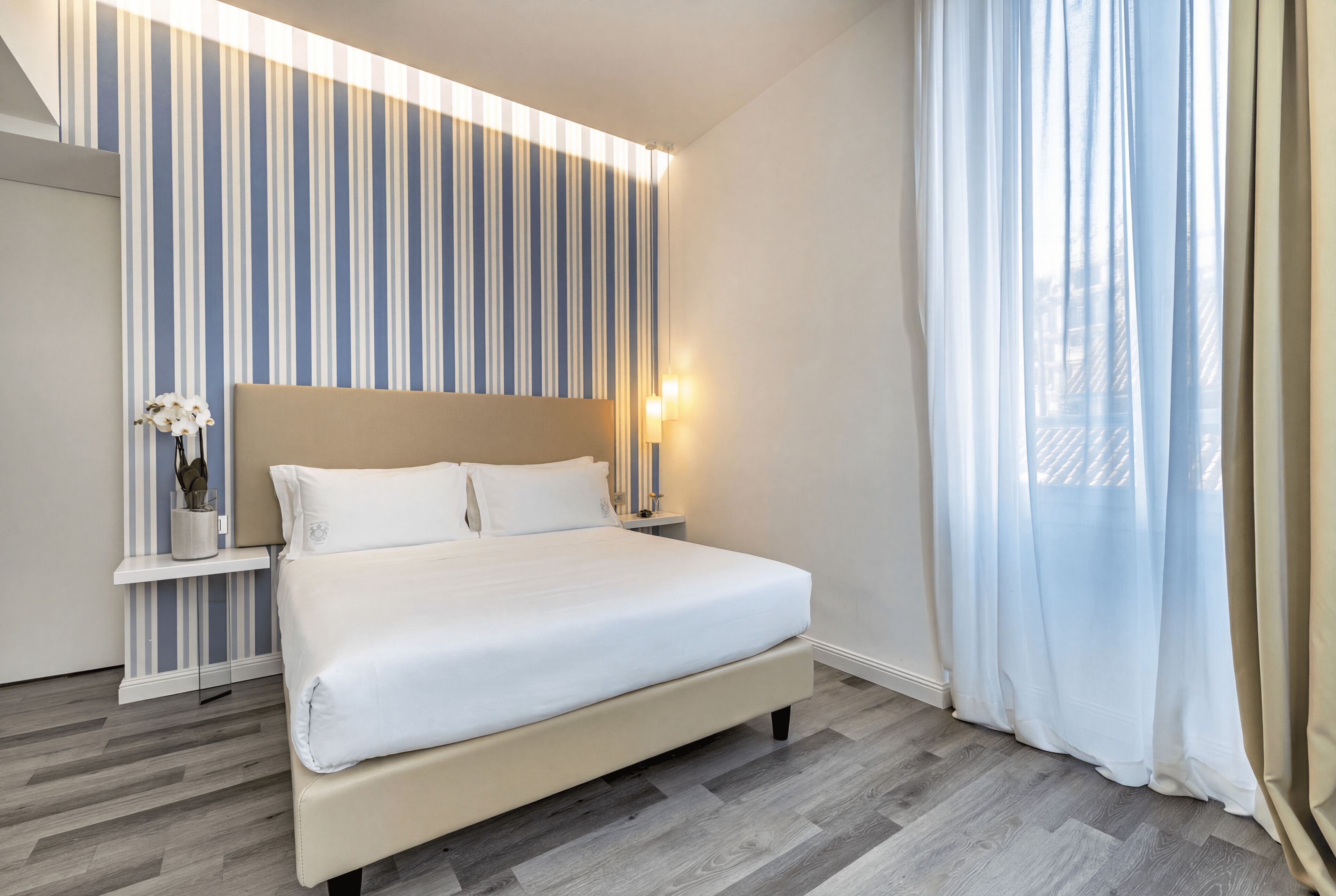 A modern hotel room with a large bed, a nightstand, a wall-mounted light, and a window with sheer curtains.