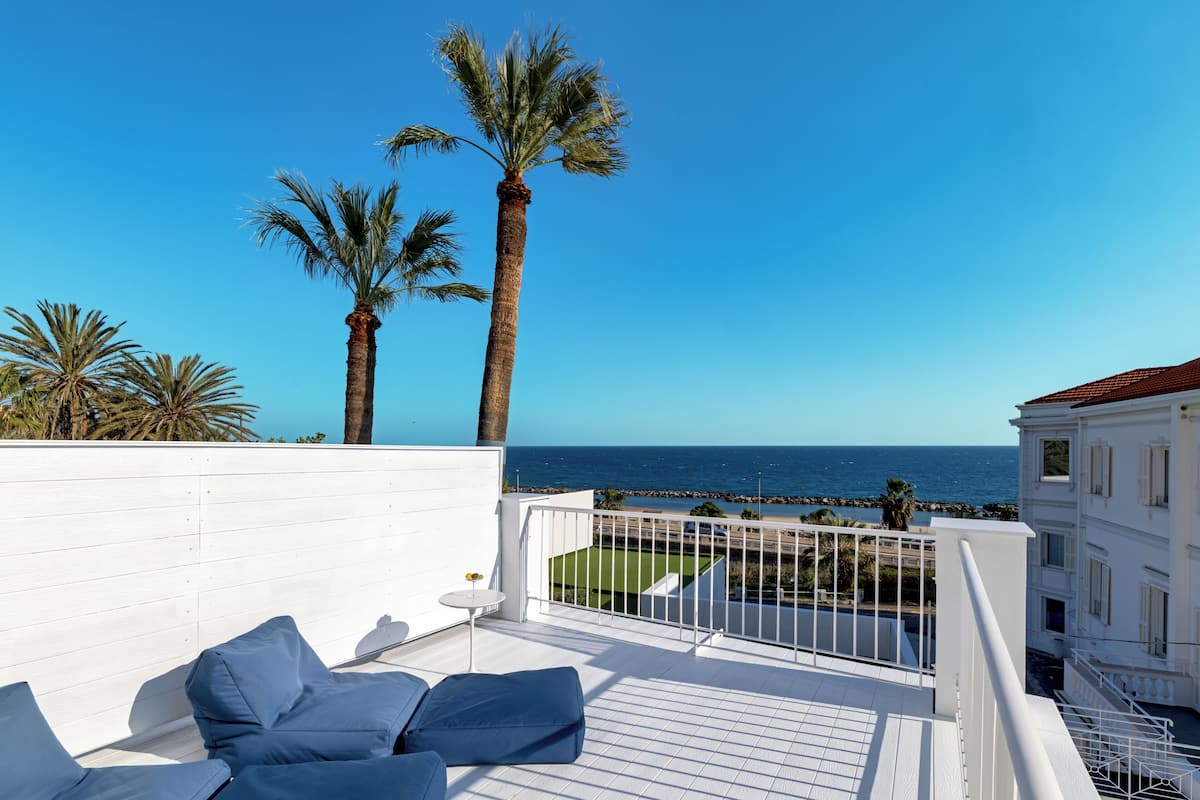A balcony with white railings, blue cushions, and a view of the sea and palm trees.