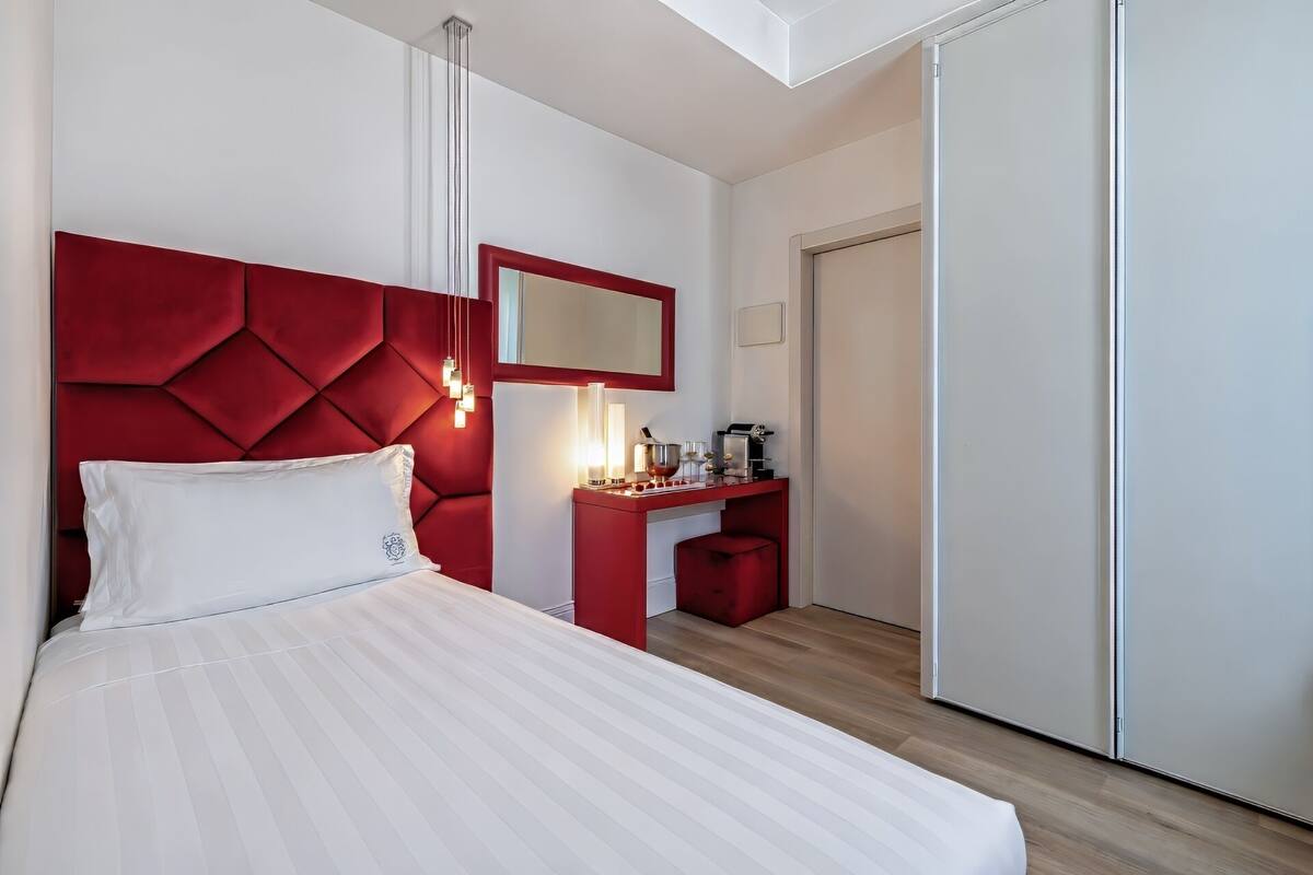 A hotel room with a bed, a red headboard, a bedside table with a coffee machine, and a mirror.