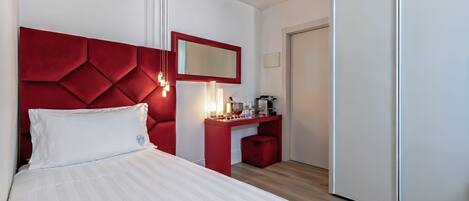 Una habitación de hotel con una cama, cabecera roja, mesita de noche con cafetera y un espejo.
