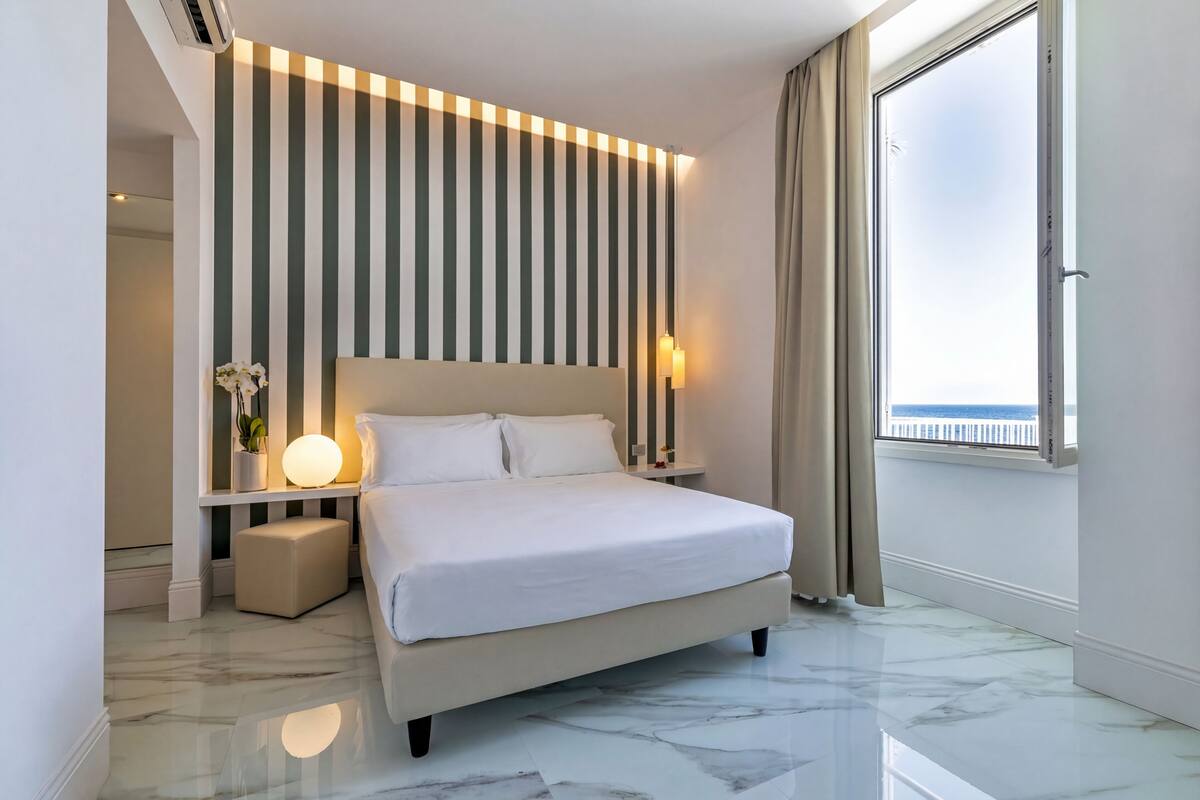 A modern hotel room with a large bed, a bedside table with a lamp, a small stool, a wall-mounted light, and a window with a view.