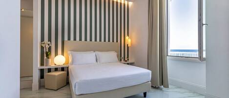 A modern hotel room with a large bed, a bedside table with a lamp, a small stool, a wall-mounted light, and a window with a view.