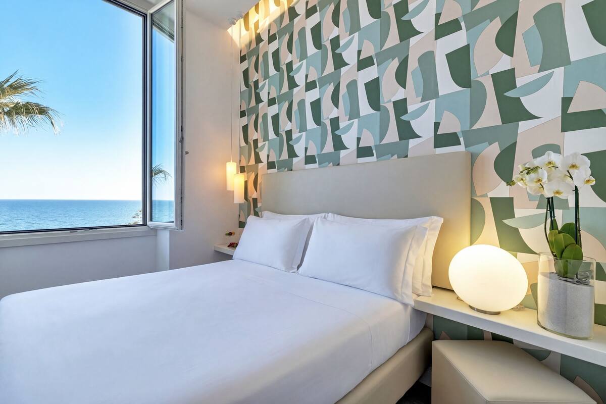 A modern hotel room with a large bed, a bedside table with a lamp, a decorative wall, and a view of the sea.