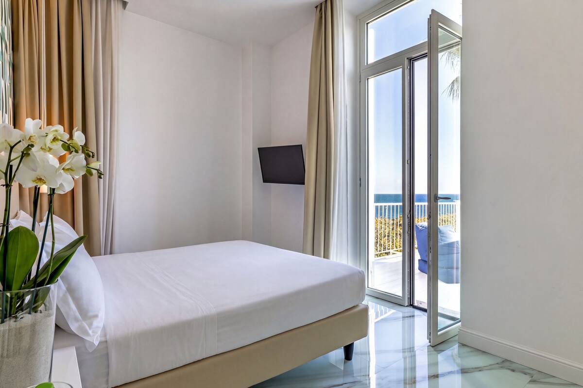 A hotel room with a bed, a TV, a sliding glass door to the balcony, and a potted plant.