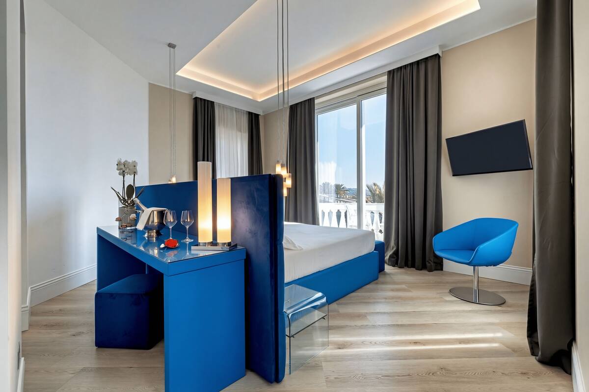 A modern hotel room with a bed, a desk with a chair, a TV mounted on the wall, and a view of the city.