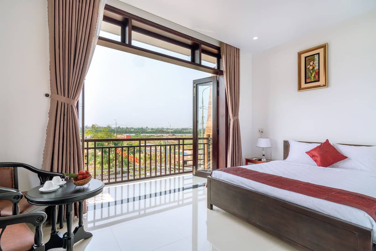 A hotel room with a bed, a table with a basket of fruit, a chair, and a balcony with a view.