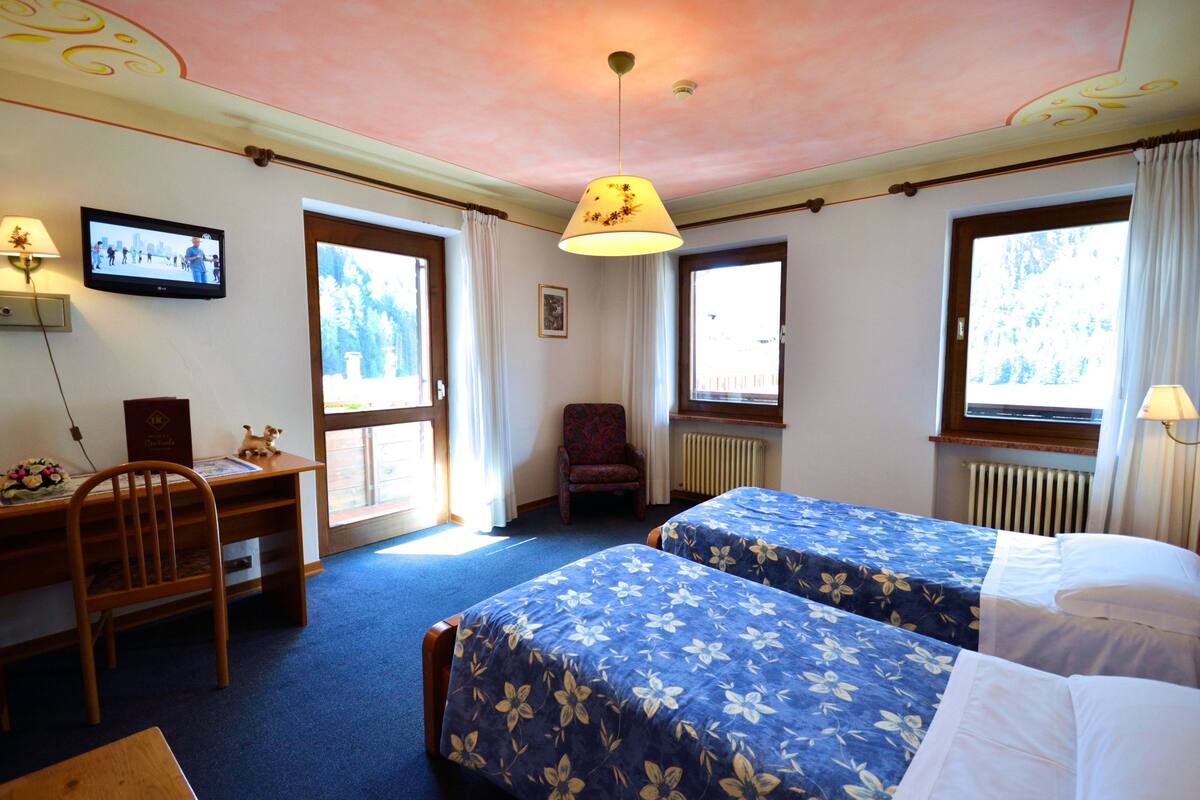 A hotel room with two beds, a desk, a chair, a television, and large windows with curtains.