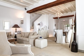 Un salón espacioso con techo alto, vigas de madera a la vista, una araña y una zona de comedor con una mesa y sillas blancas.