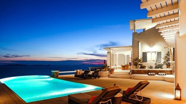 A poolside area with lounge chairs, a small table, and a view of the sea at dusk. - Villa Sunshine (Mykonos)