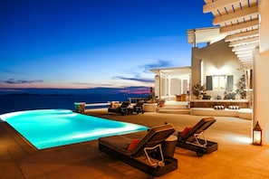 Zona junto a la piscina con sillones reclinables, una mesita y vistas al mar al atardecer.
