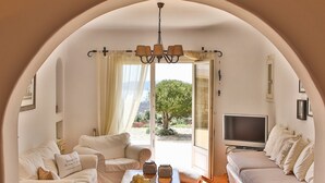 A cozy living room with a sofa, a coffee table, and a TV. There is a large window with curtains, and a tree is visible outside. - Villa Sunshine (Mykonos)
