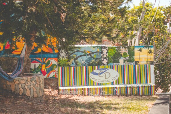A colorful mural with tropical birds and palm trees, a stone bench, and a fence with the text "Cima Dorada Villa".