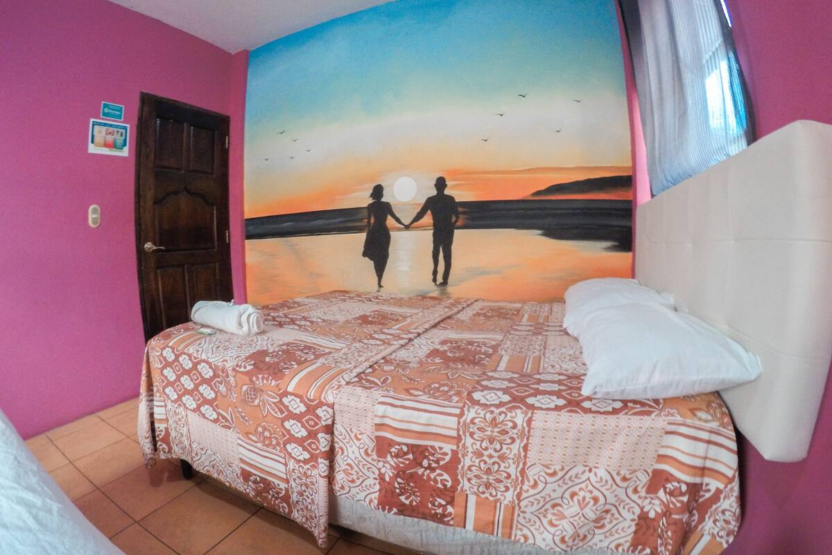 Une chambre avec un lit, une fresque murale représentant un coucher de soleil, une porte et un sol carrelé.