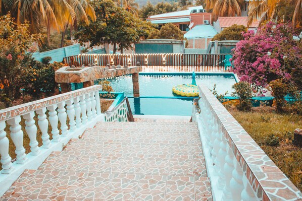 Un área de piscina con baldosas, un borde de piedra, un flotador inflable pequeño y vistas de edificios y árboles circundantes.