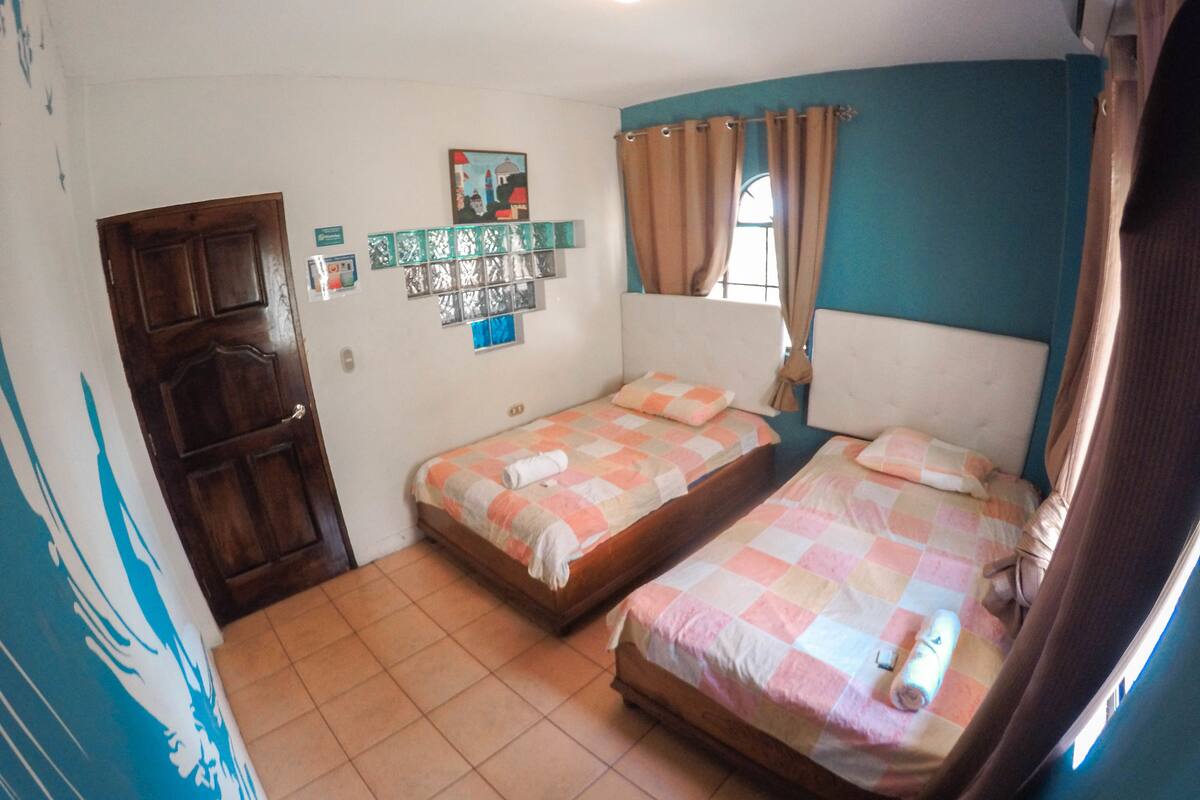 Une chambre avec deux lits simples, une porte en bois, une petite fenêtre avec des rideaux et une étagère fixée au mur, sur laquelle sont disposés divers objets.