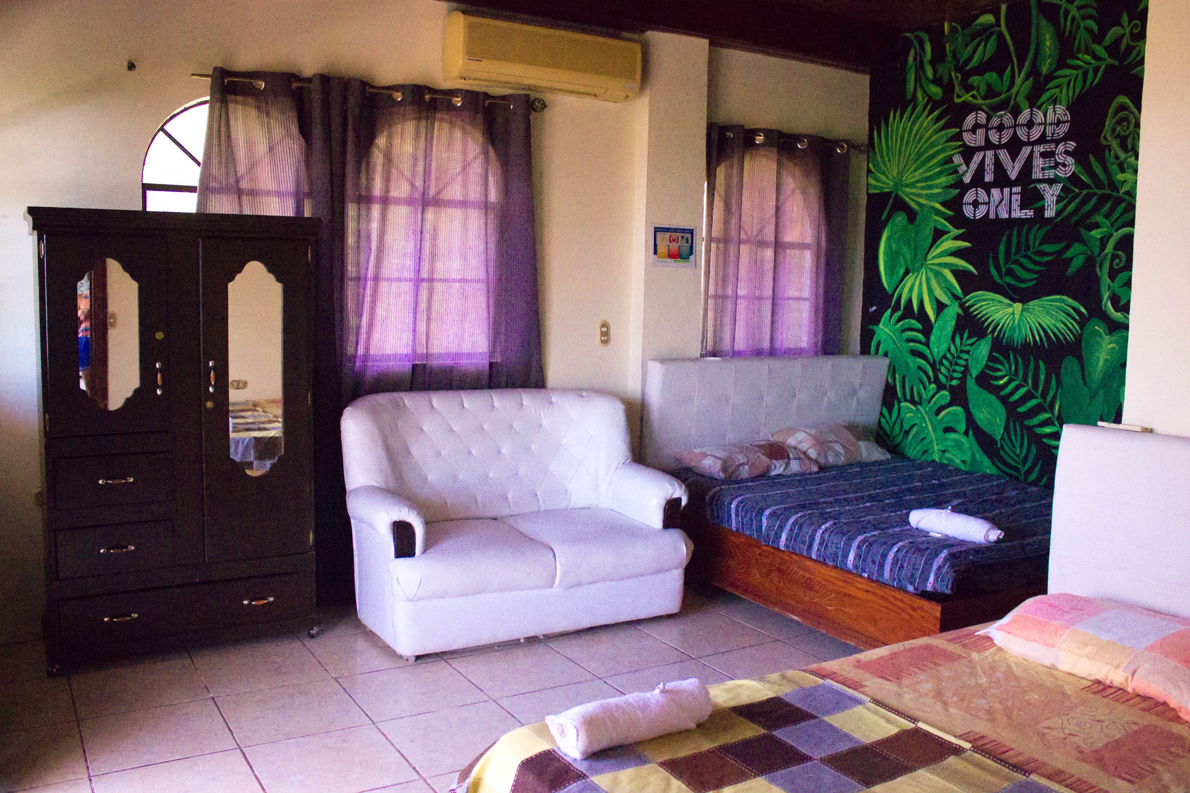 A bedroom with a bed, a white armchair, a wooden wardrobe, and a wall with a tropical plant mural.