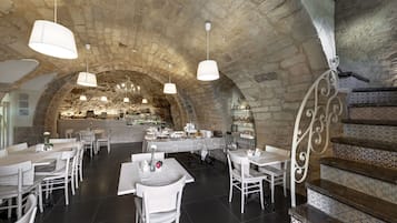 Un restaurante con arcos de piedra, mobiliario blanco y una zona de comedor central con cocina al fondo.