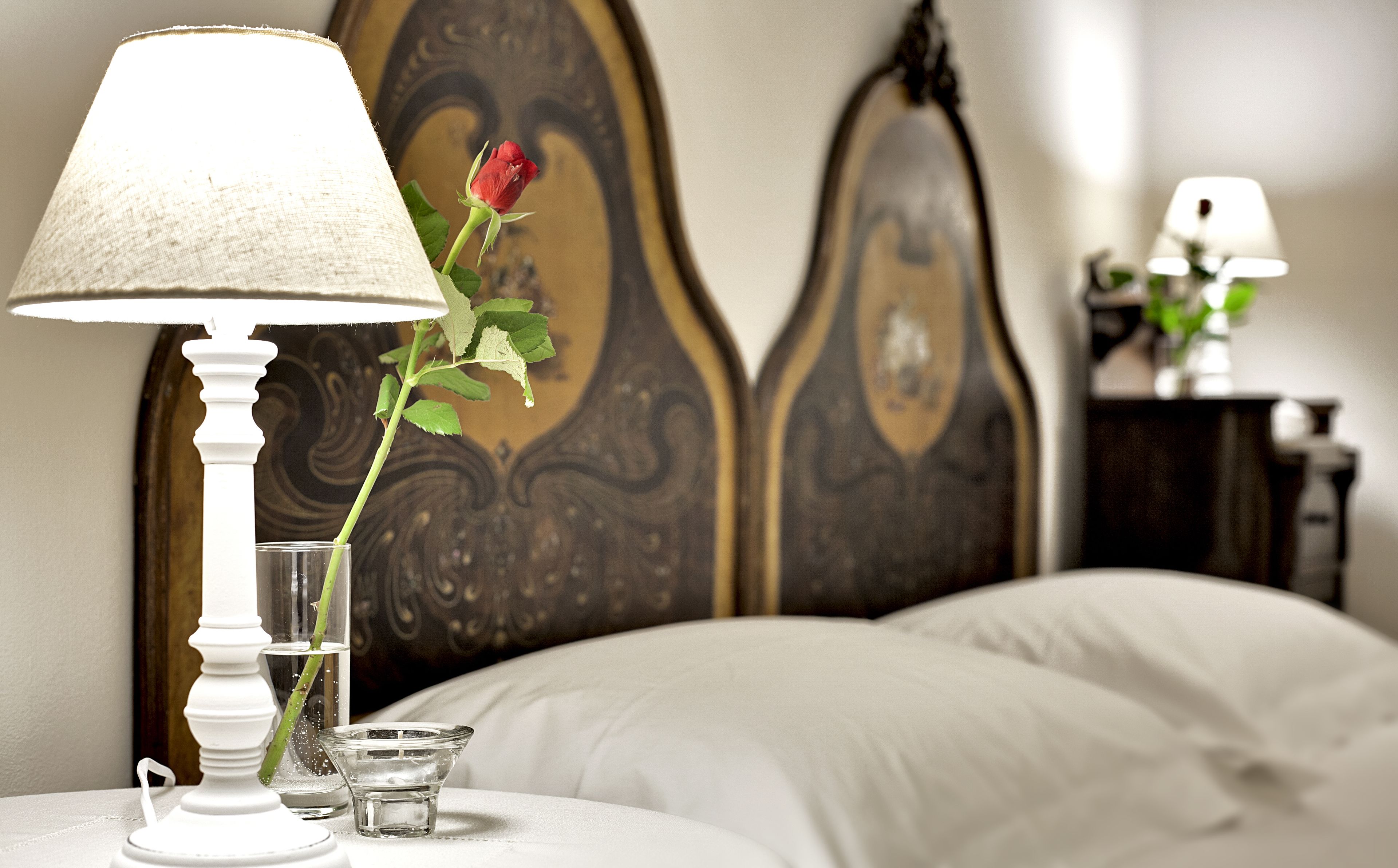 A neatly made bed with white linens, a white bedside table with a lamp and a glass of water, and a single red rose.