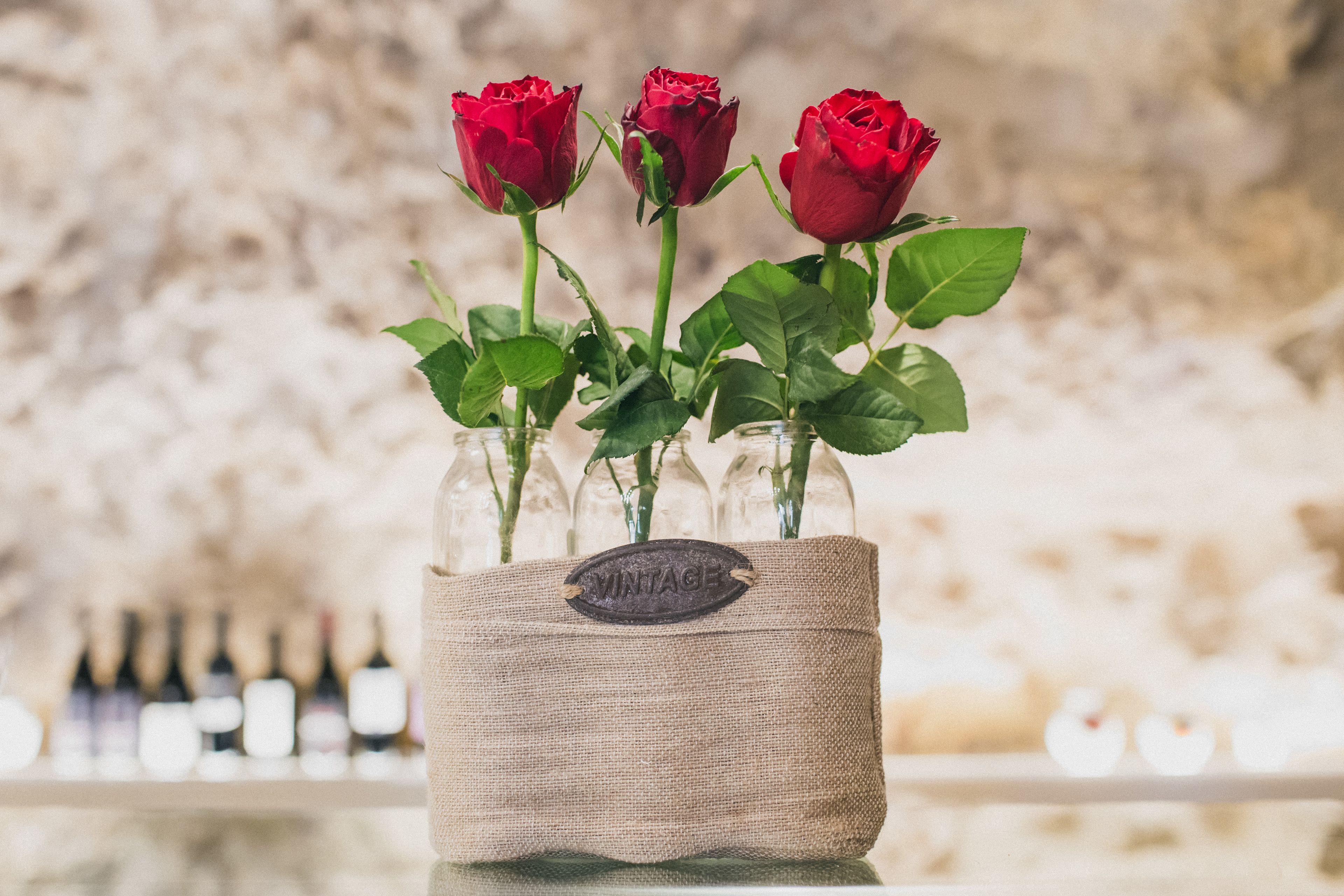 Un vase contenant trois roses rouges, placé dans un récipient en toile de jute portant l’étiquette « VINTAGE », le tout posé sur une surface réfléchissante.