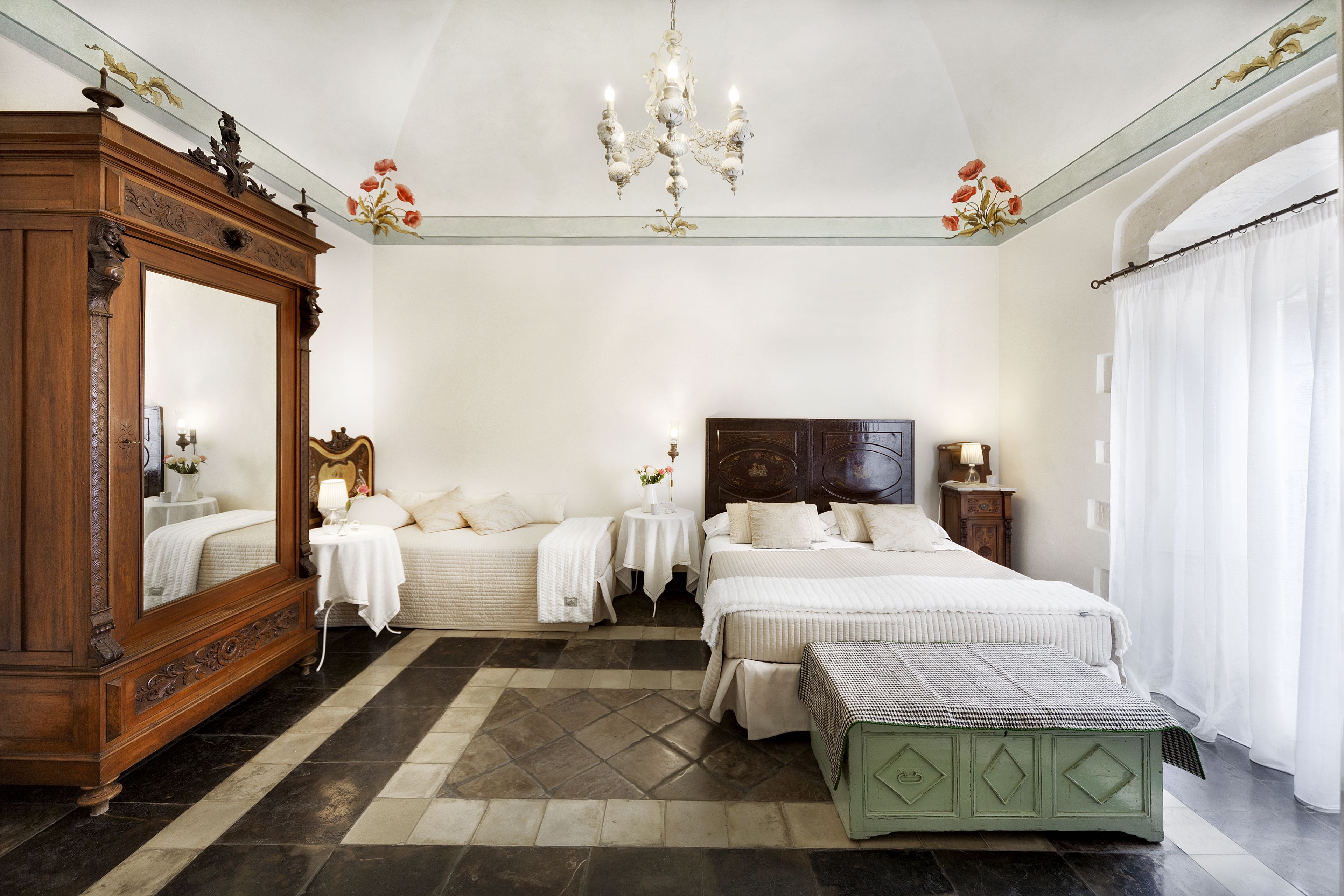 Une chambre avec un grand miroir, un lustre, un lit avec une literie blanche et un banc vert.