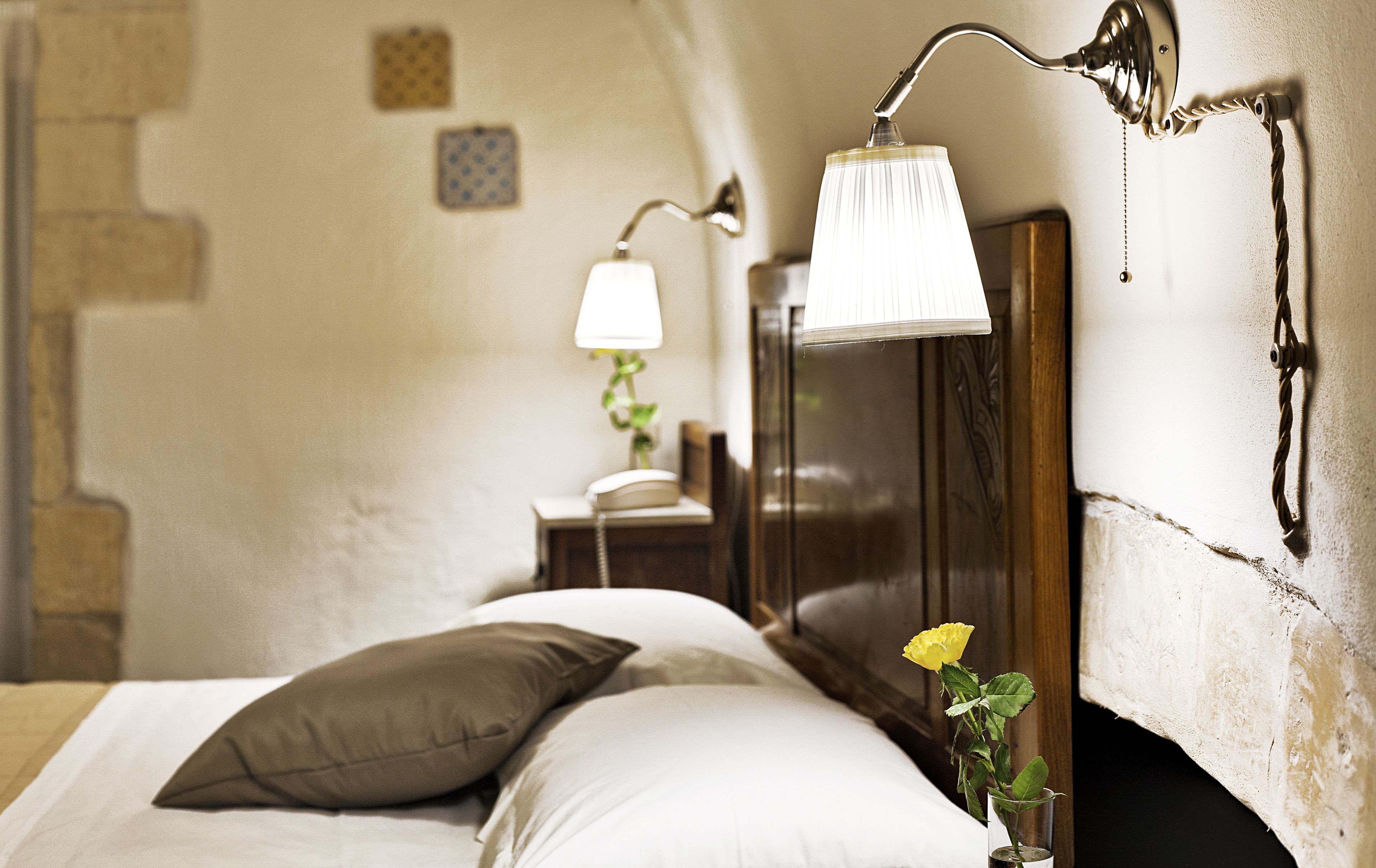 A bedroom with a bed, pillows, a bedside lamp, a nightstand, a telephone, and a wall-mounted lamp.