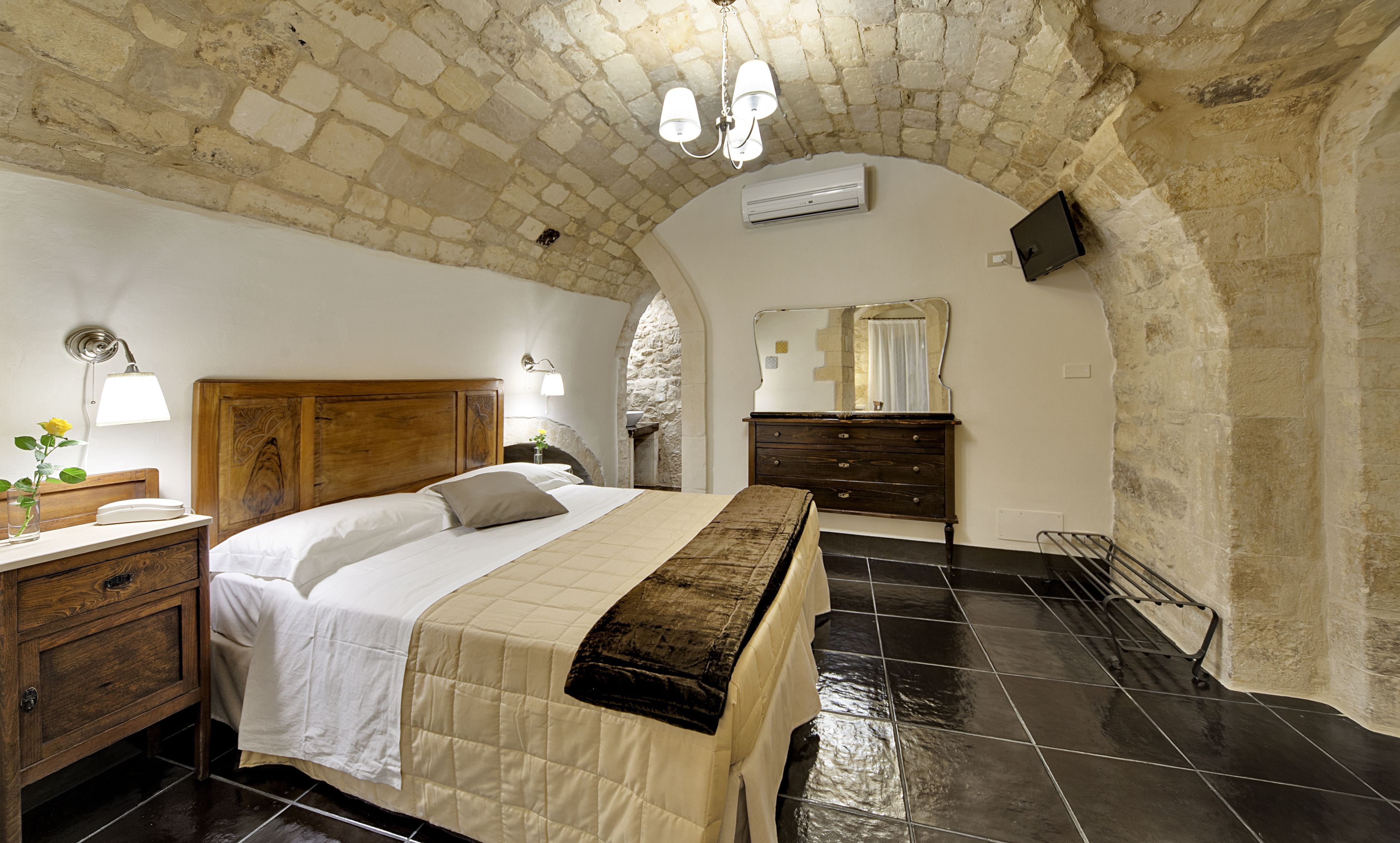 A bedroom with a stone wall, a wooden bed, a dresser, and a TV mounted on the wall.