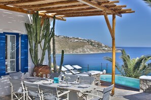 A terrace with a table set for dining, overlooking a coastal landscape with a pool and lounge chairs. - Villa Athena (Mykonos)