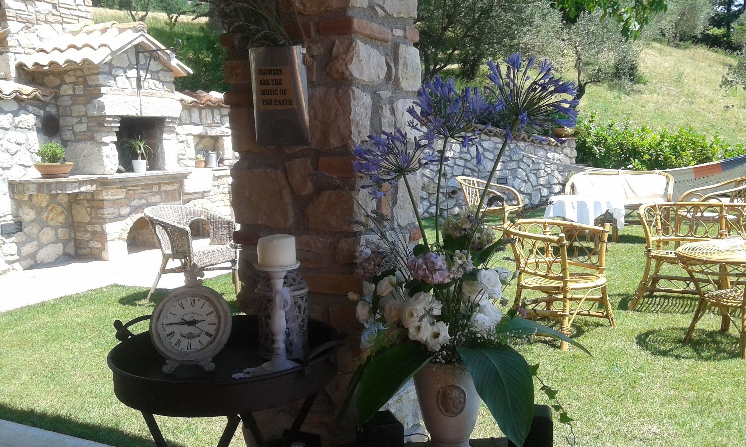 Un muro de piedra con una placa, sillas de mimbre, una mesa con un reloj y un arreglo floral.