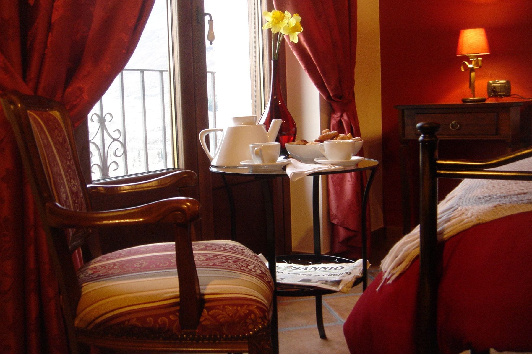 Ein Hotelzimmer mit Holzstuhl, einem kleinen Tisch mit Teekanne, Tassen und Gebäck, einem Bett mit roter Bettwäsche und einer Lampe auf einem Nachttisch.