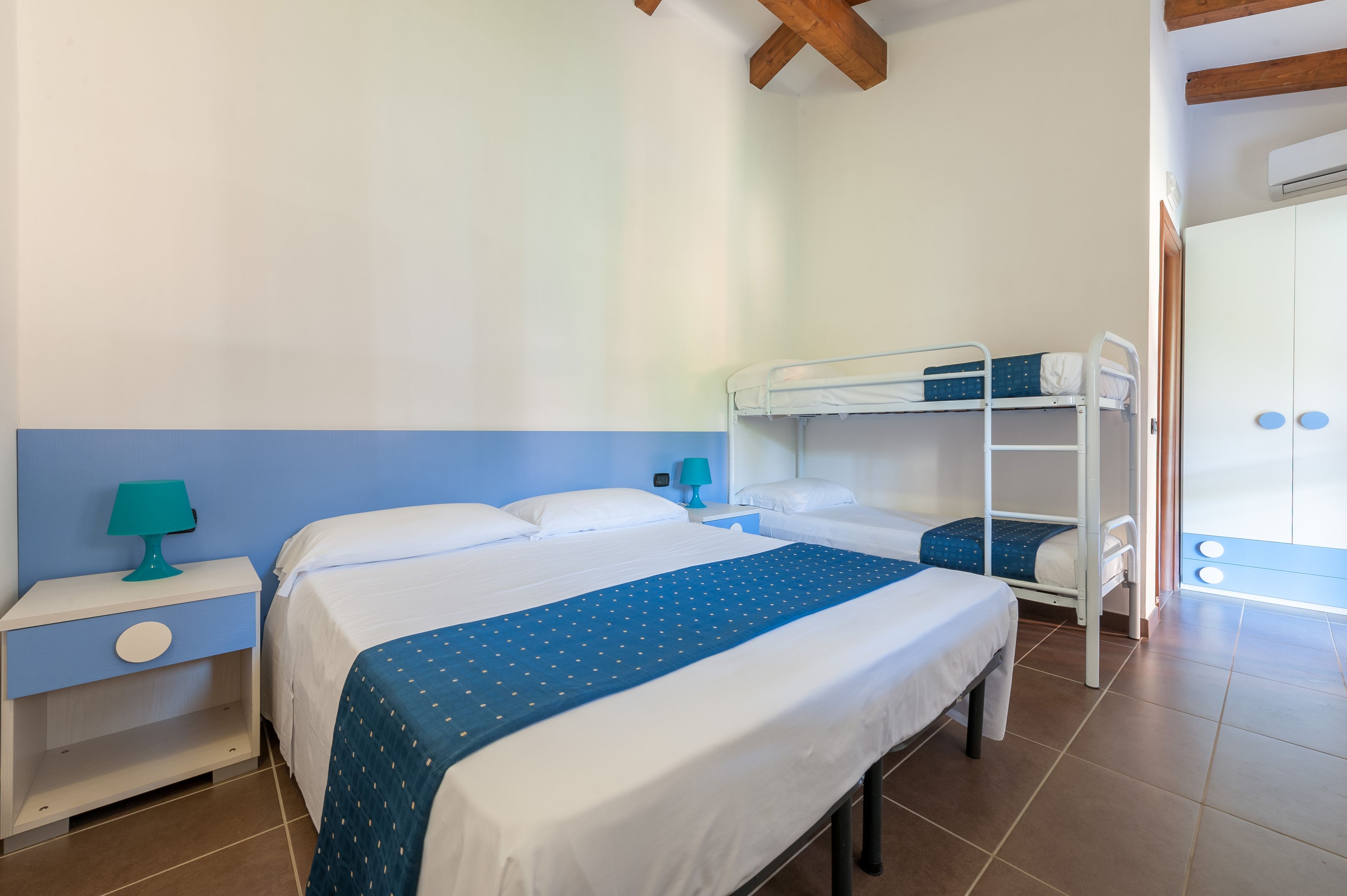 Una habitación de hotel con una cama grande, una cama nido más pequeña, una mesita de noche con lámpara y un armario.