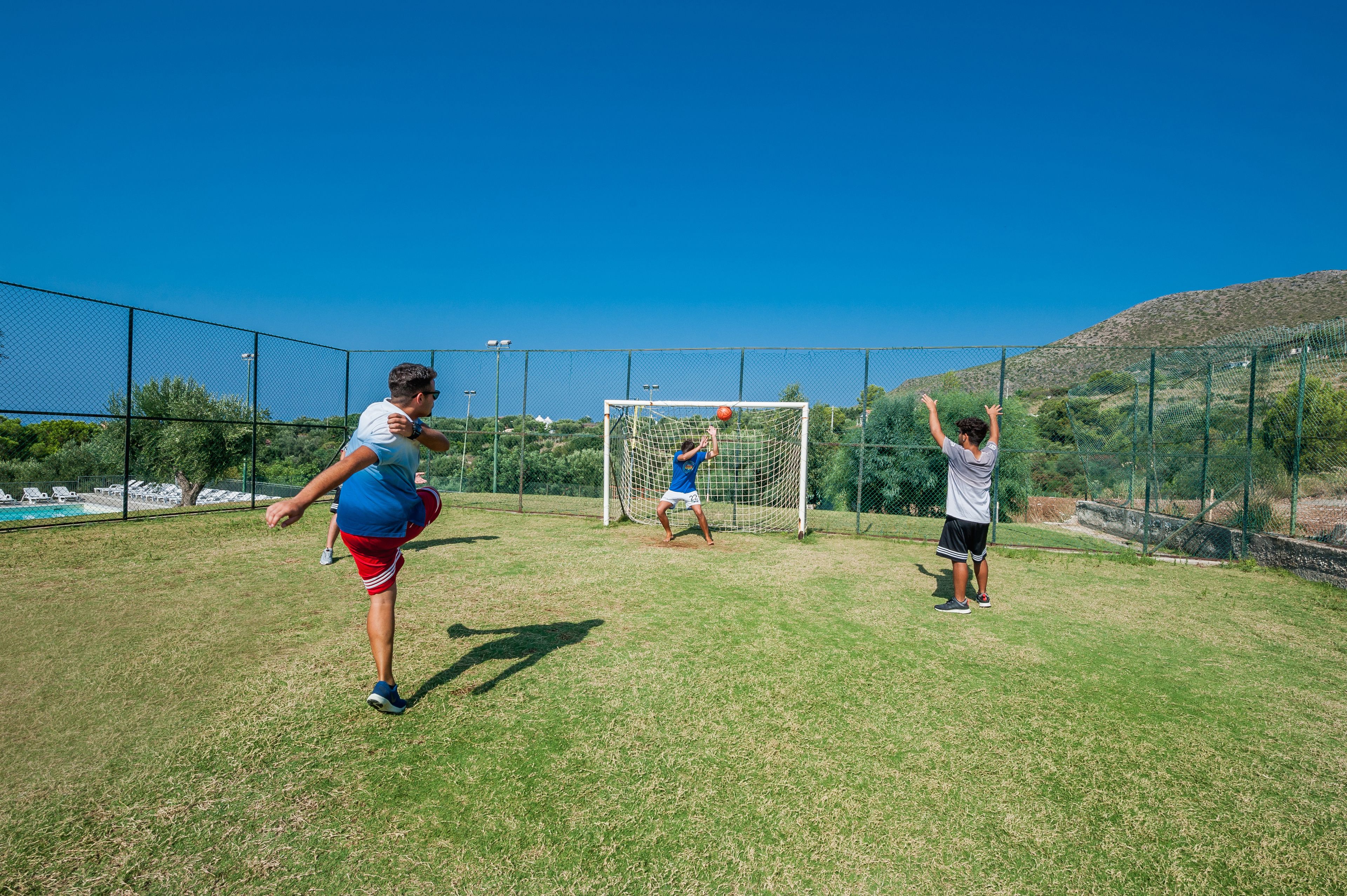A soccer field with a goalpost, surrounded by a fence and a hilly landscape in the background.