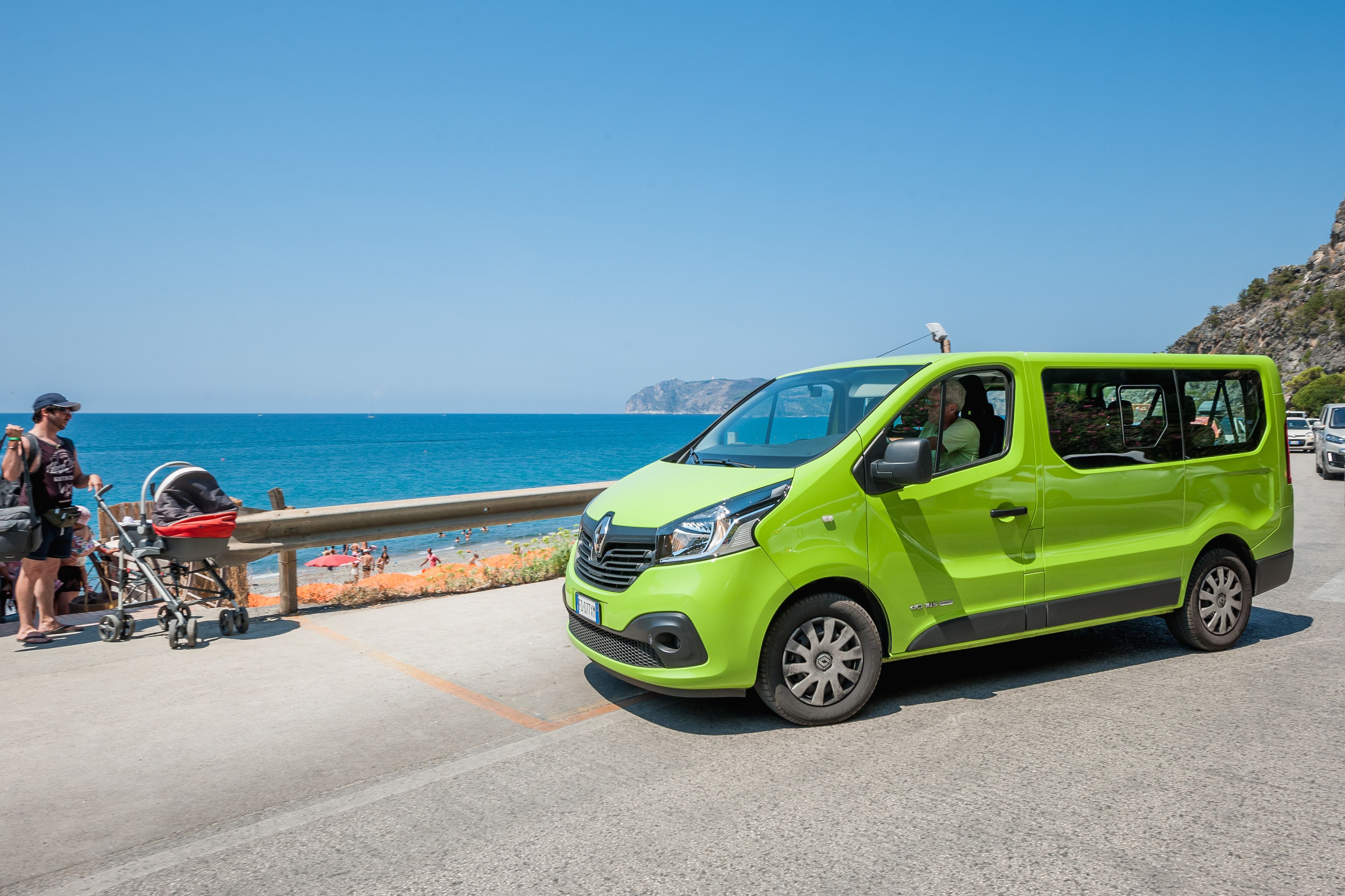 Una minifurgoneta verde brillante estacionada al borde de una carretera costera, con una vista despejada del mar y acantilados rocosos al fondo.
