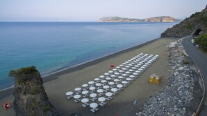 Una playa con hileras de sombrillas y mesas, un litoral rocoso y una carretera que serpentea a lo largo de la costa.
