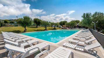Una piscina con sillas de descanso blancas dispuestas a su alrededor, rodeada de árboles y un cielo despejado.