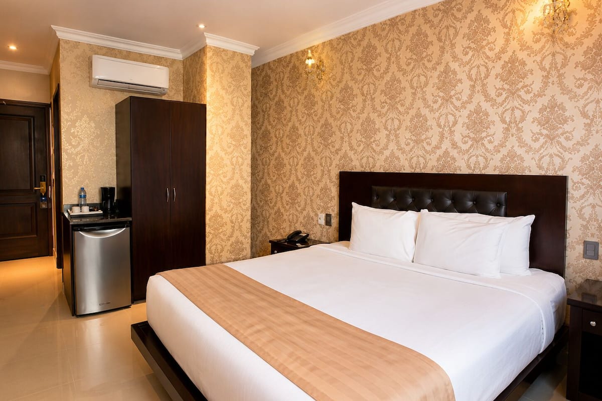 A hotel room with a large bed, a wardrobe, a desk, a chair, a trash bin, and an air conditioning unit.
