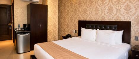 A hotel room with a large bed, a wardrobe, a desk, a chair, a trash bin, and an air conditioning unit.