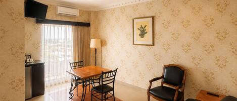 A hotel room with a dining table, chairs, a sofa, a TV, an air conditioning unit, and a framed picture on the wall.