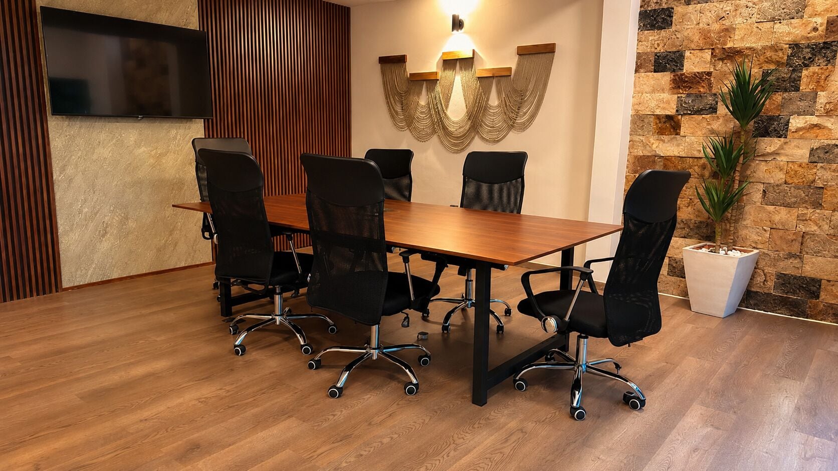 Un salón de conferencias con una mesa de madera, sillas negras, un televisor, una lámpara fijada en la pared y una pared de piedra.