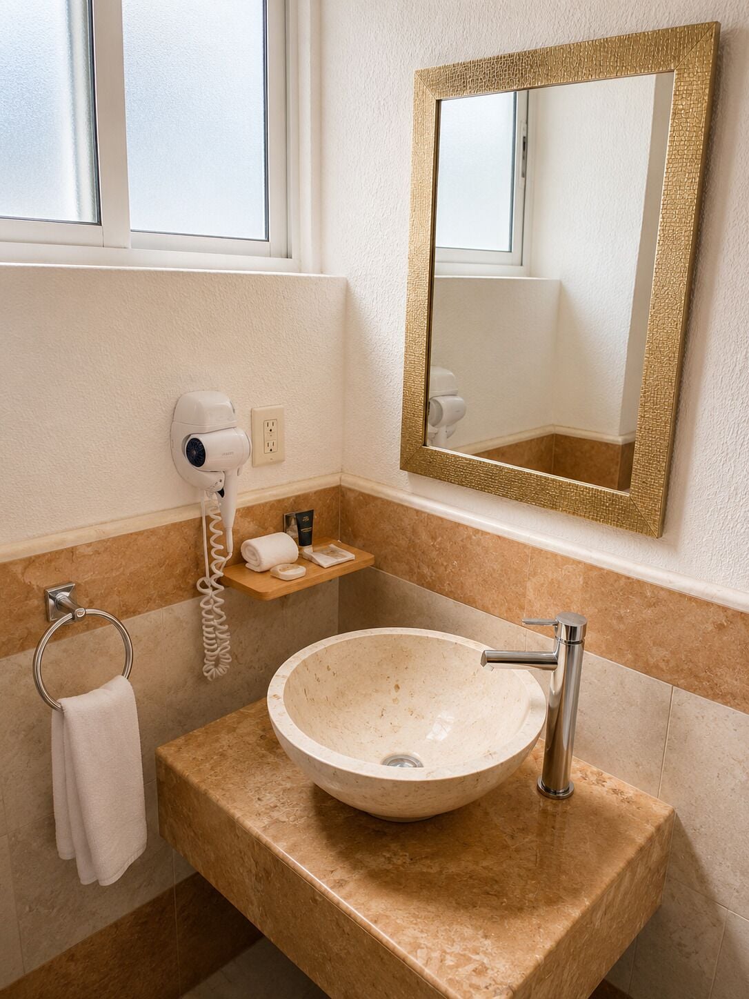 A bathroom with a beige sink, a gold-framed mirror, a towel rack, and a hairdryer on a wall-mounted shelf.