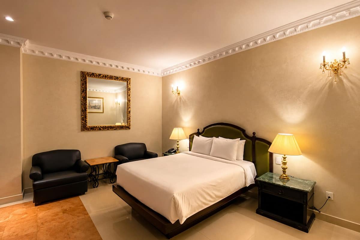 A hotel room with a large bed, two chairs, a small table, a mirror, and a chandelier.