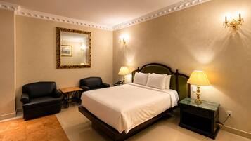 A hotel room with a large bed, two chairs, a small table, a mirror, and a chandelier.