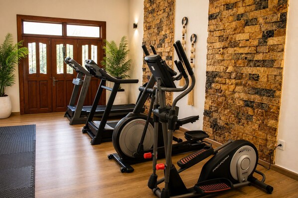 A home gym with treadmills, an elliptical, and a stationary bike, featuring a wooden floor, a stone wall, and a door with glass panels. - Hotel Palacio Maya (Mérida)