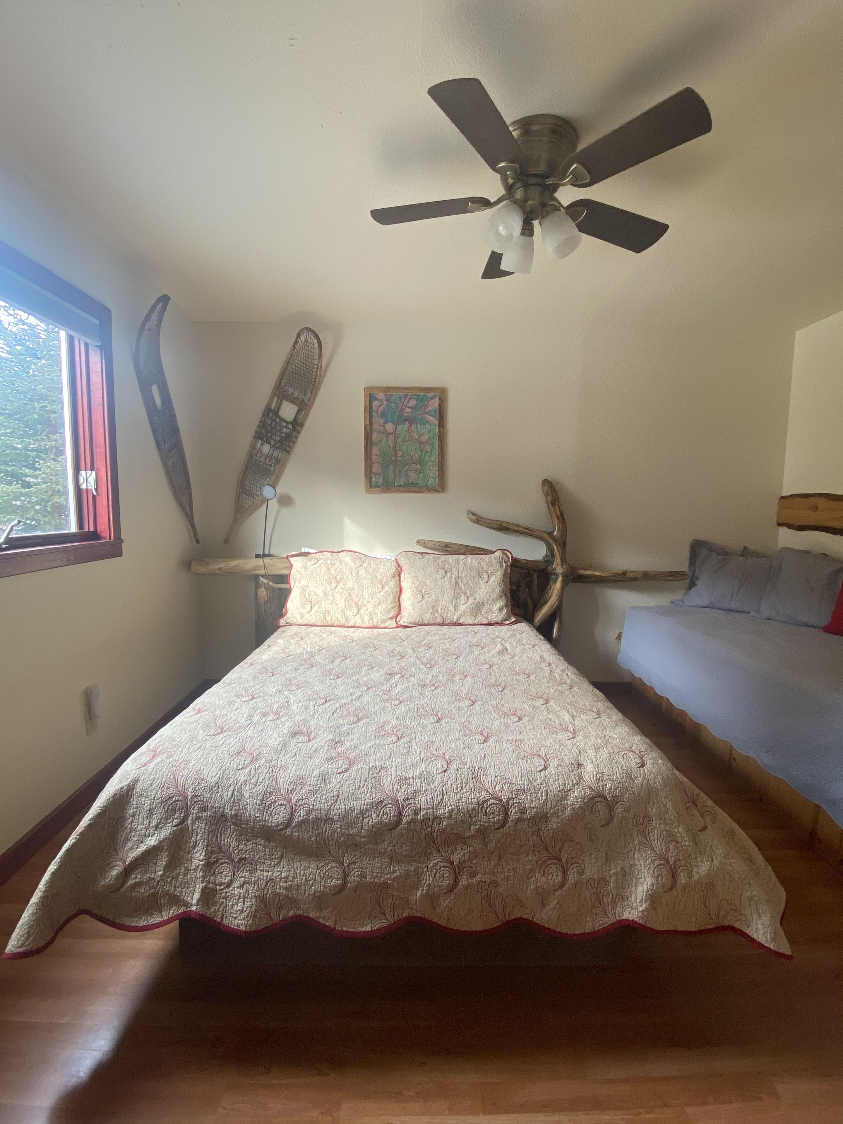 A bedroom with a bed, a ceiling fan, a window, and a wall-mounted artwork.